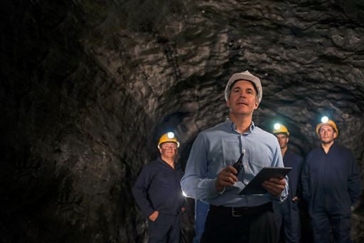 homem com capacete de proteçao segurando uma prancheta dentro de um túnel de mineração com 3 homens ao fundo com capacete de proteção com laternas