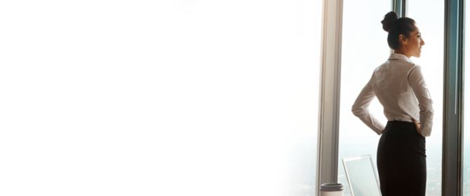 em primeiro plano mostra uma mesa com papéis e uma xicara de café. em segundo plano uma executiva de saia longa preta, blusa branca e cabelos em formato de coque olhando através da janela de vidro