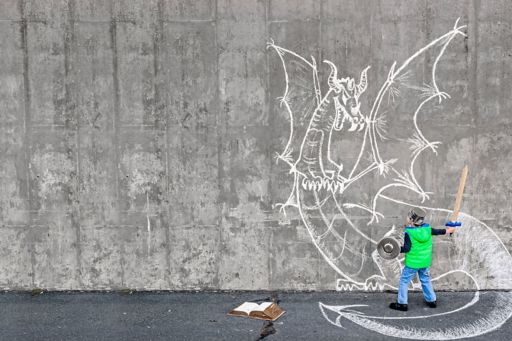 Child holding sword and facing wall where dragon is drawn with chalk , book