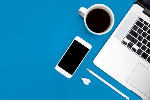 Smartphone, Kaffeetasse, Stift, kleiner Papierflieger neben Laptop auf blauem Hintergrund