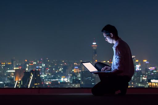 Man working on laptop late night sitting at roof, city lights background
