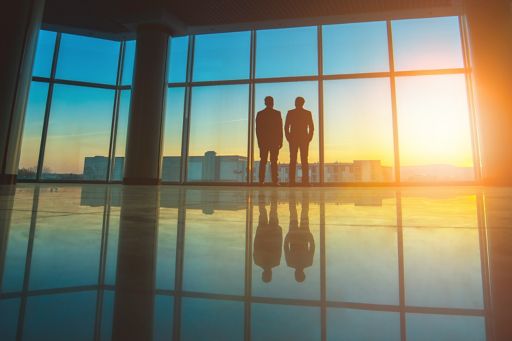 Two businessmen standing at office glass window at sunset