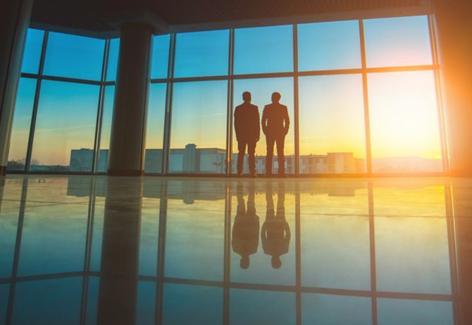 Two businessmen standing at office glass window at sunset