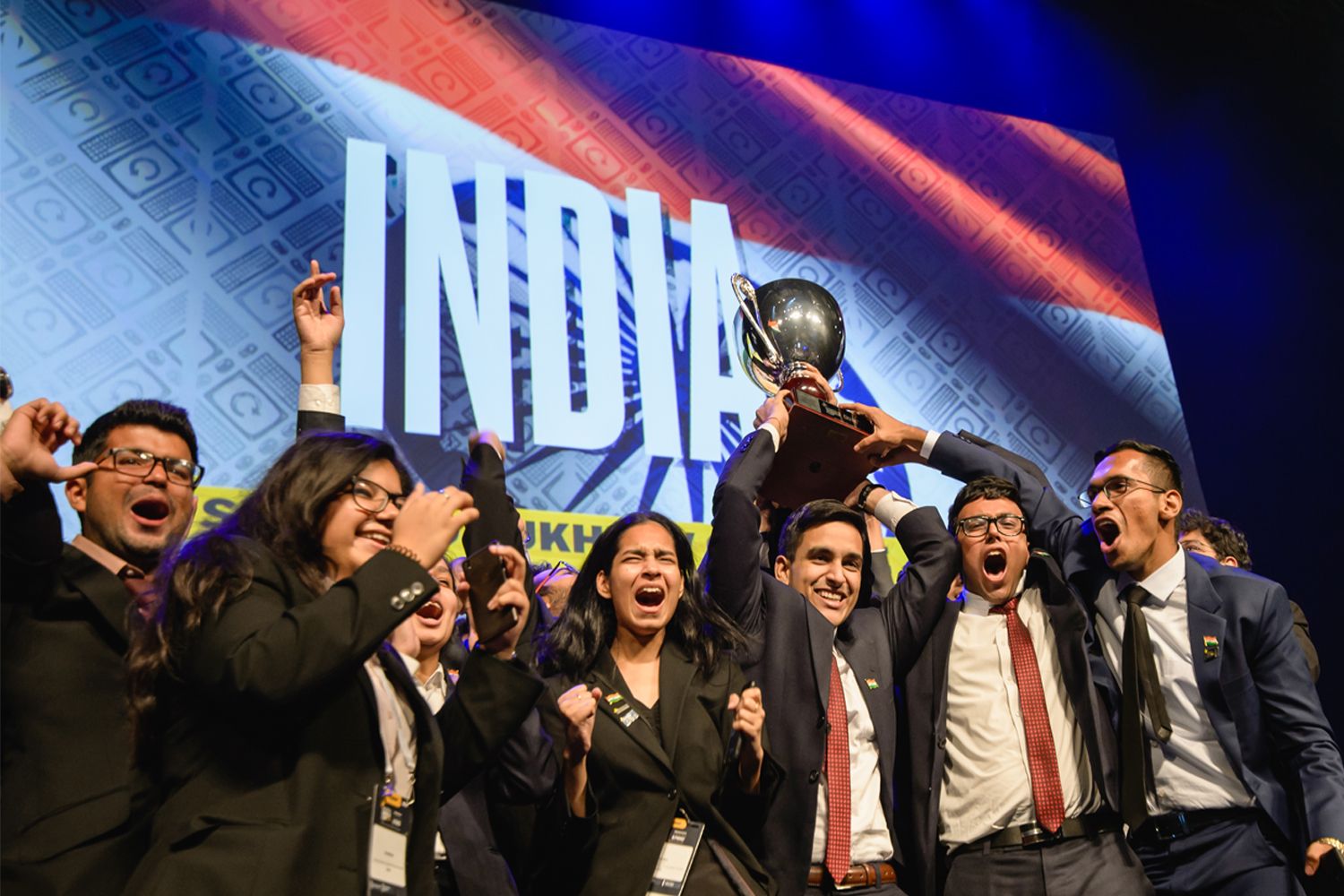 Students wearing business suits and holding trophy in front of India banner