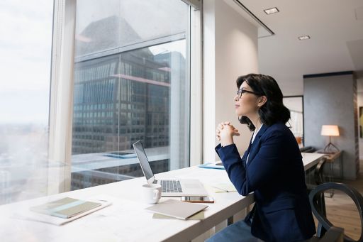 Woman sitting and gazing out of office with laptop in-front